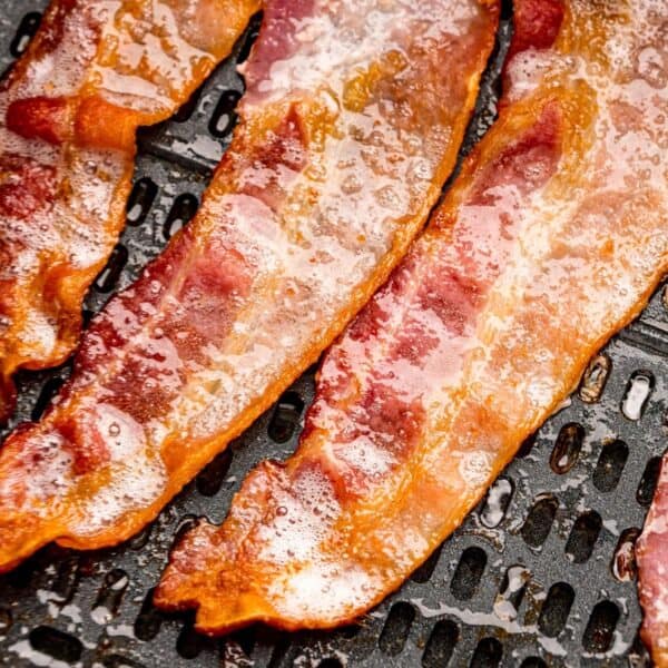 Three strips of cooked bacon with visible grease, resting on a black slotted tray, show how to use an air fryer for perfectly crisp results.