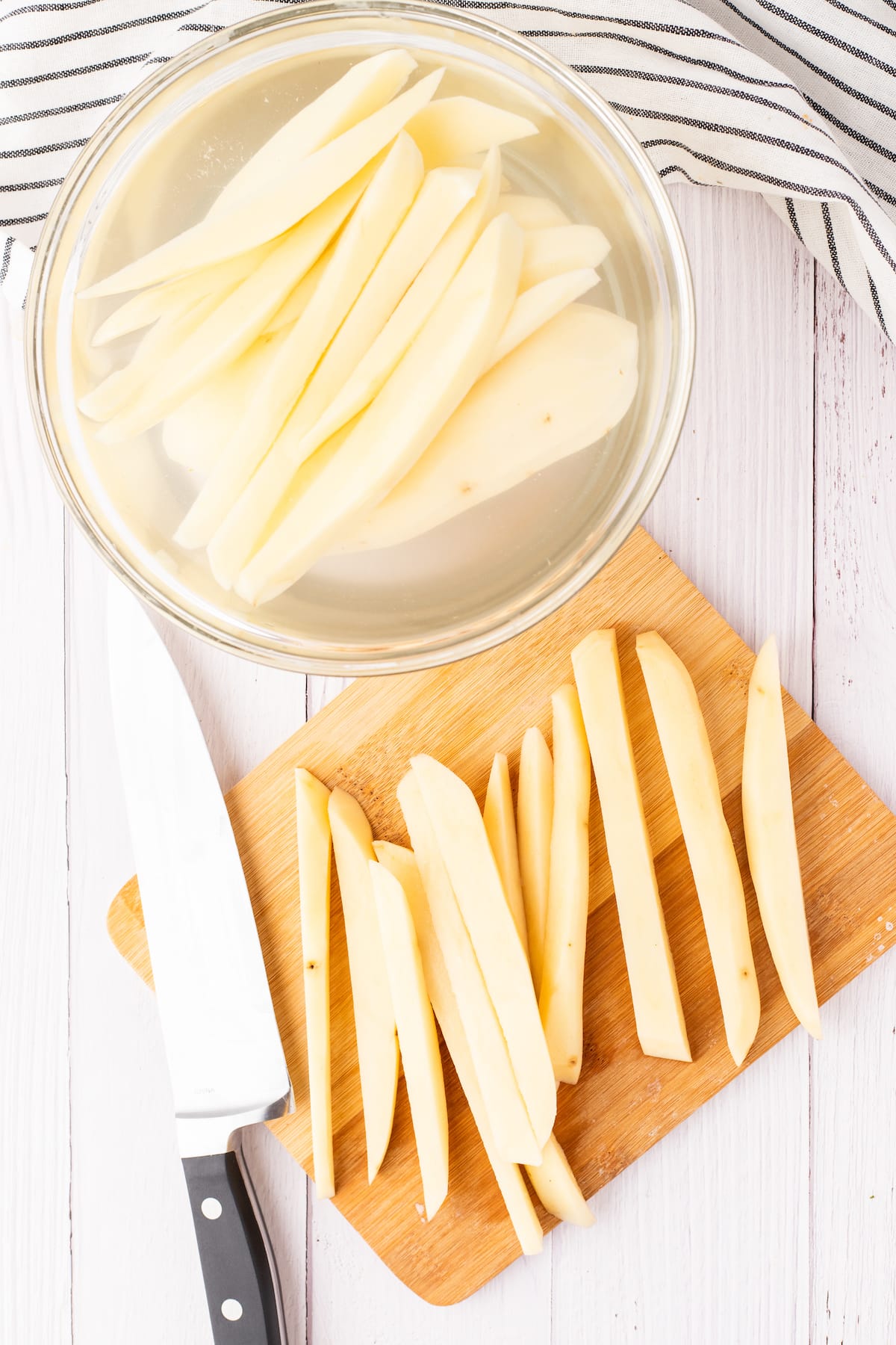 potato cut into fries on a cutting board next to fries soaking in water