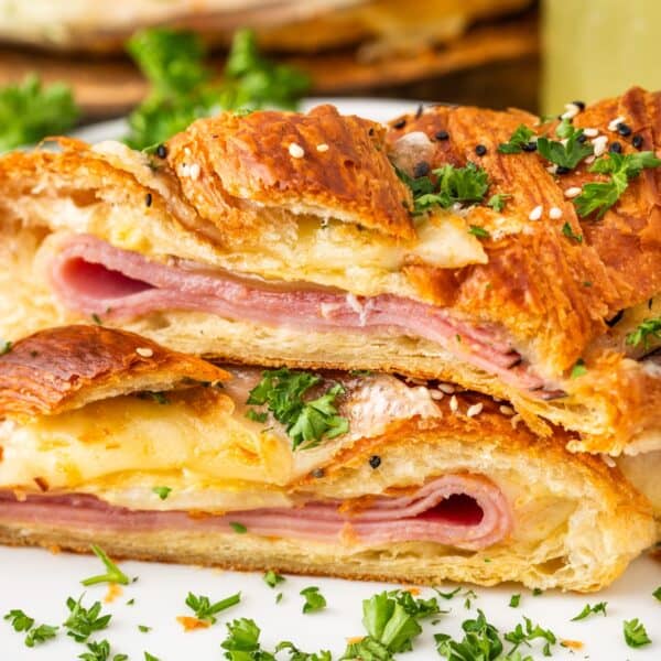 Two slices of braided puff pastry filled with ham and melted cheese, reminiscent of baked ham and cheese croissants, garnished with chopped parsley and sesame seeds, served on a white plate.