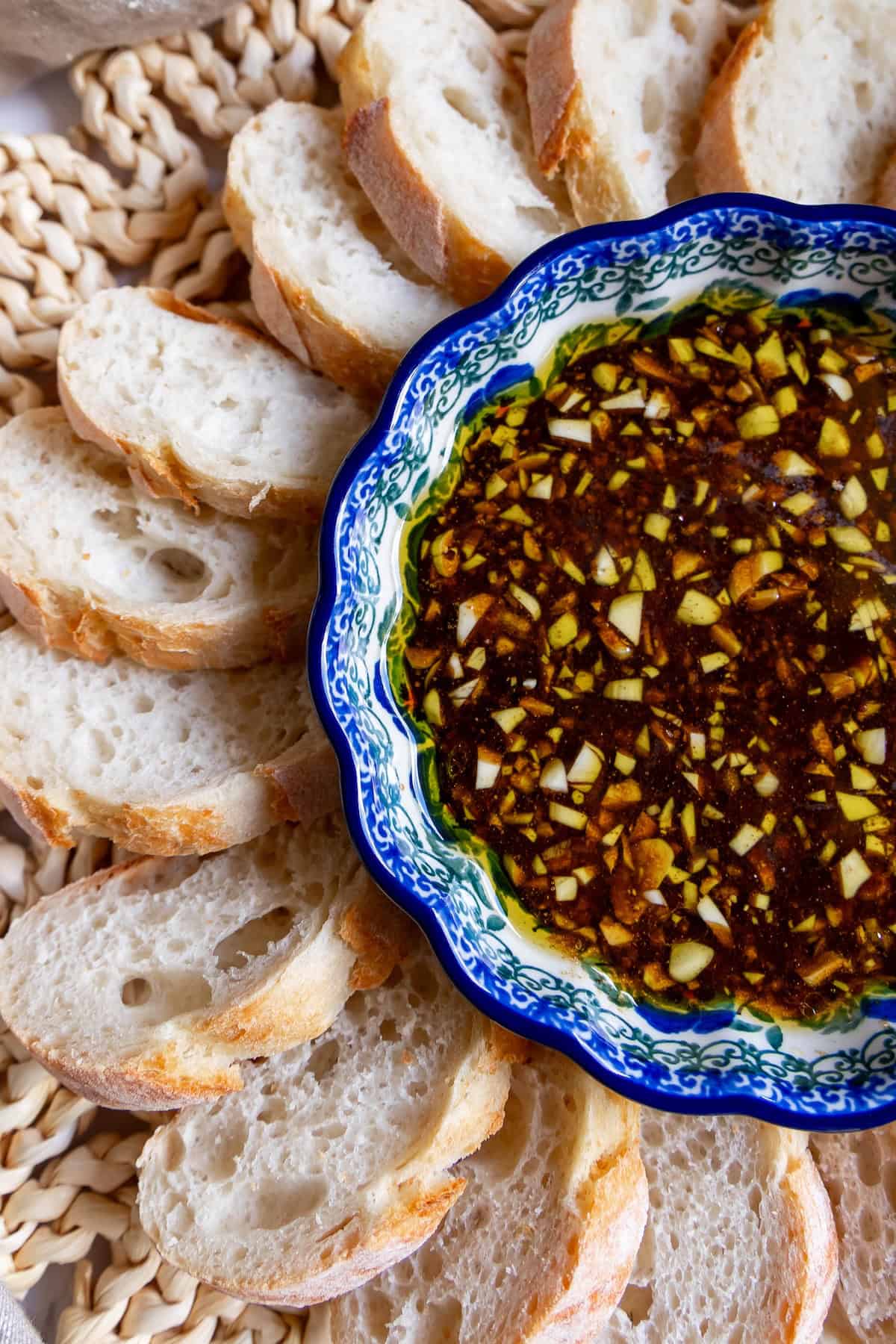 A decorative bowl filled with chopped garlic and herbs in balsamic vinaigrette, surrounded by sliced pieces of crusty bread on a woven mat.