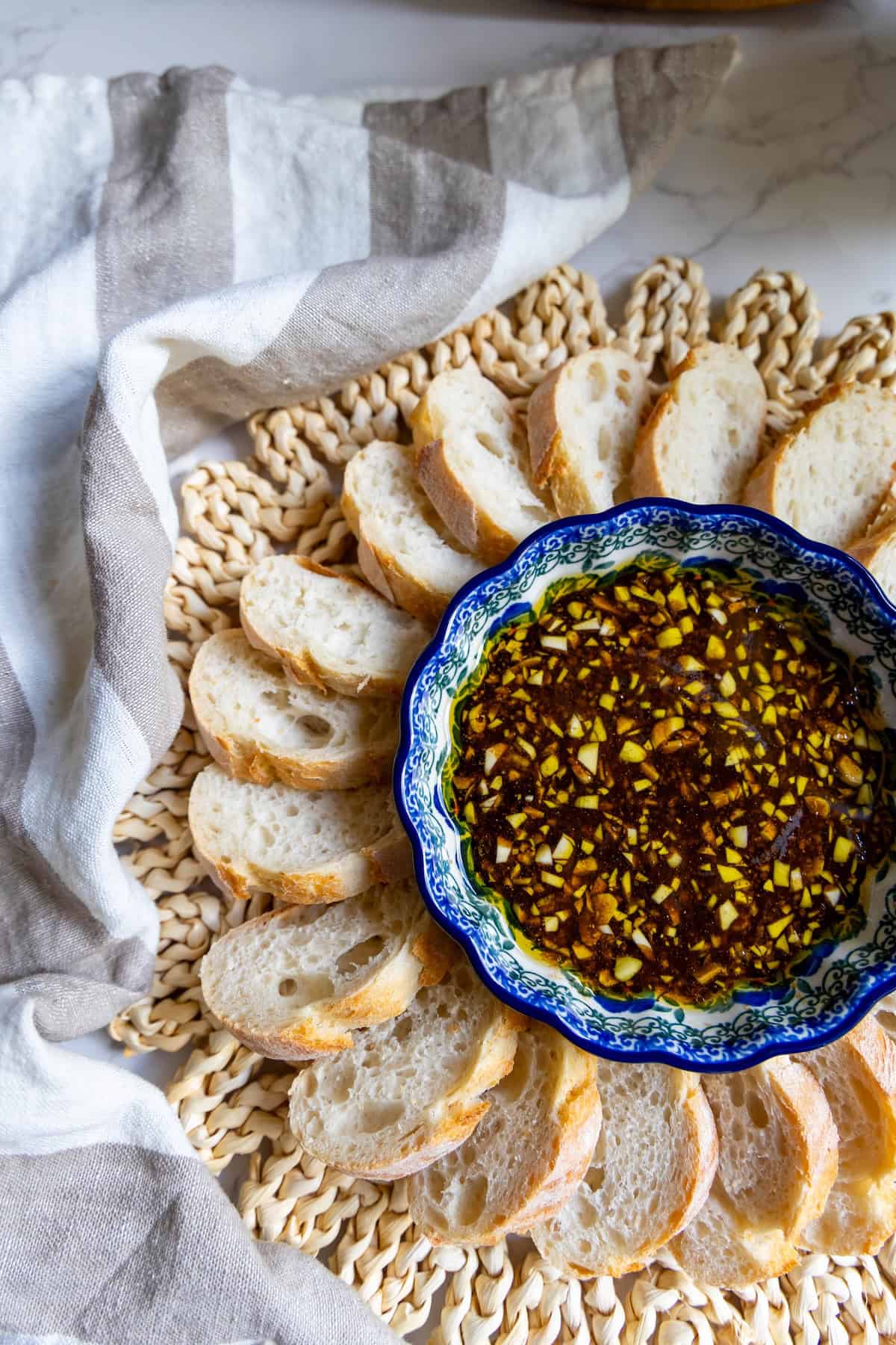 A bowl of chopped garlic and herb oil dip, reminiscent of a rustic balsamic vinaigrette, is surrounded by slices of baguette on a woven placemat with a striped cloth nearby.