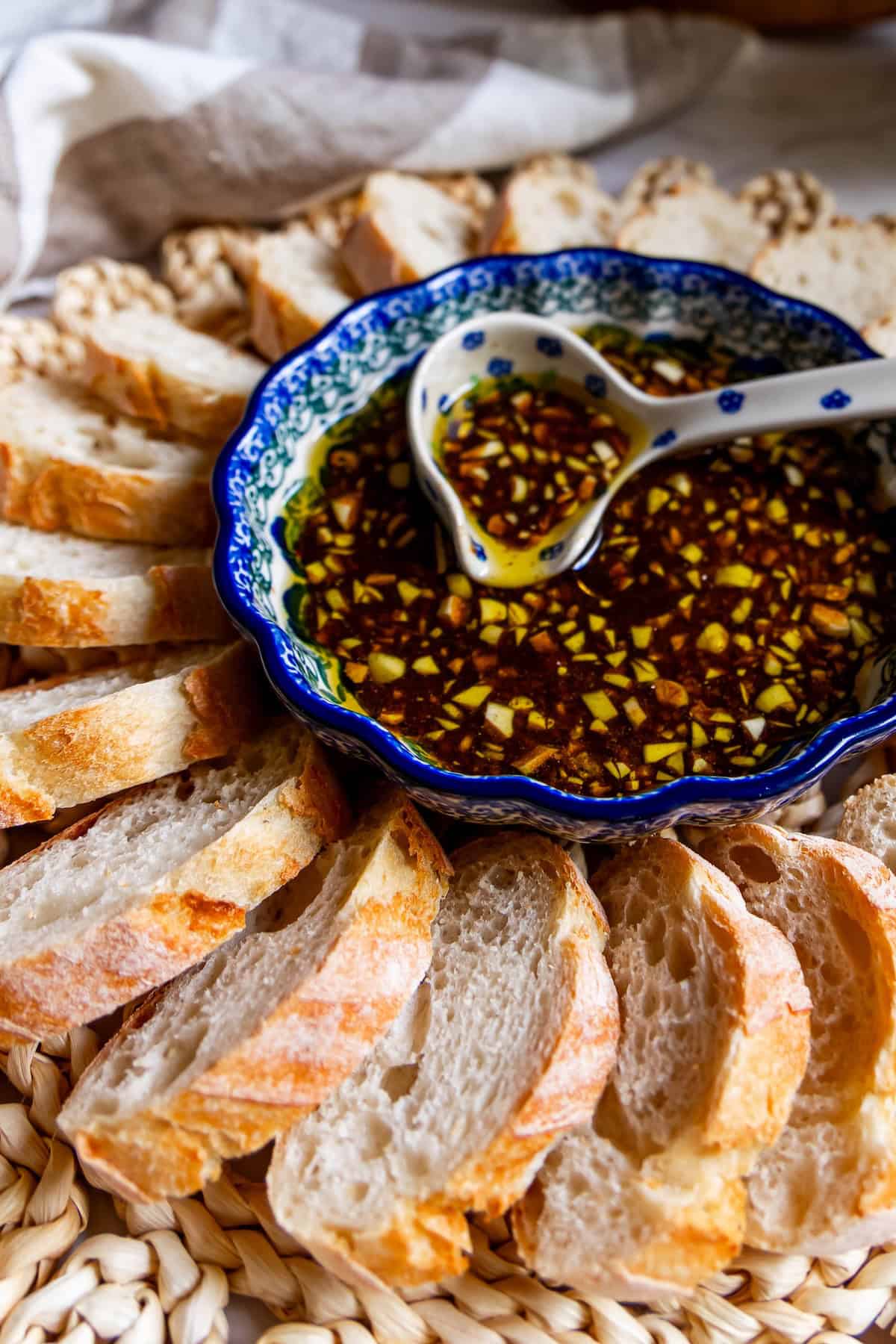 A bowl of oil and herb dip with a hint of balsamic vinaigrette sits with a spoon, surrounded by slices of crusty bread arranged in a circular pattern on a woven mat.