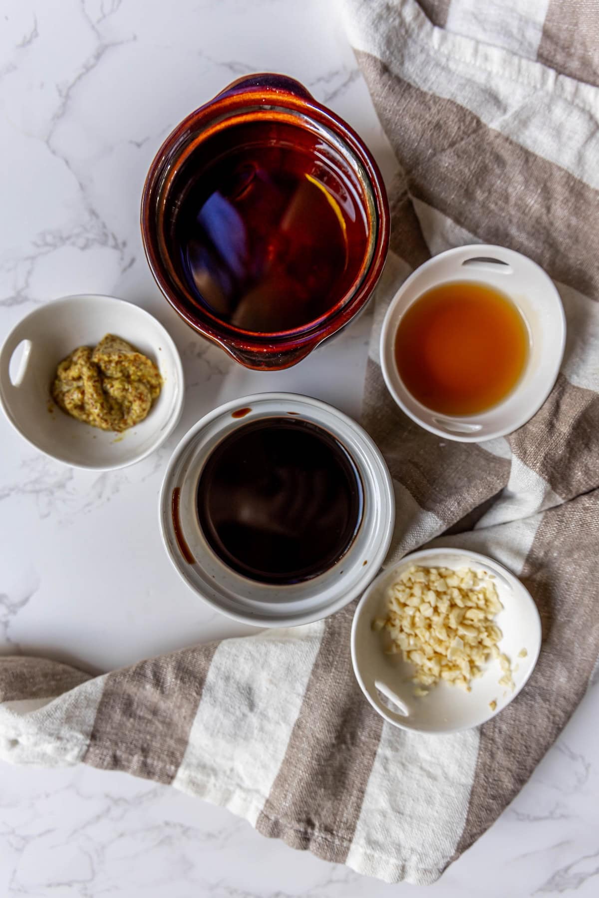 Five small bowls on a striped cloth contain ingredients for a vinaigrette: one with brown liquid, one with mustard, one with light brown liquid, one with chopped garlic, and one with dark balsamic.