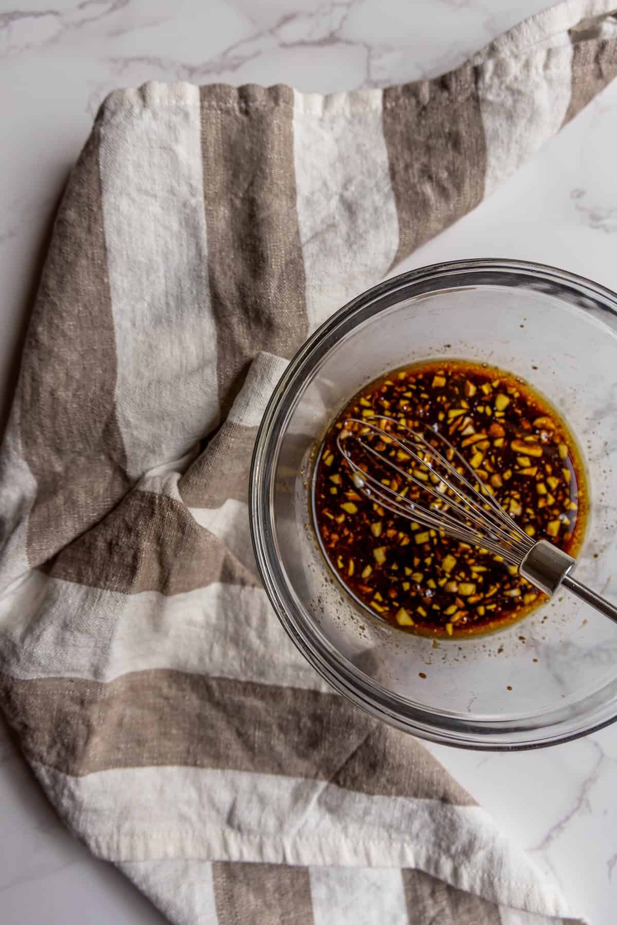 A glass bowl with a metal whisk mixing a dark balsamic vinaigrette with chopped ingredients, placed on a beige and white striped cloth on a marble surface.