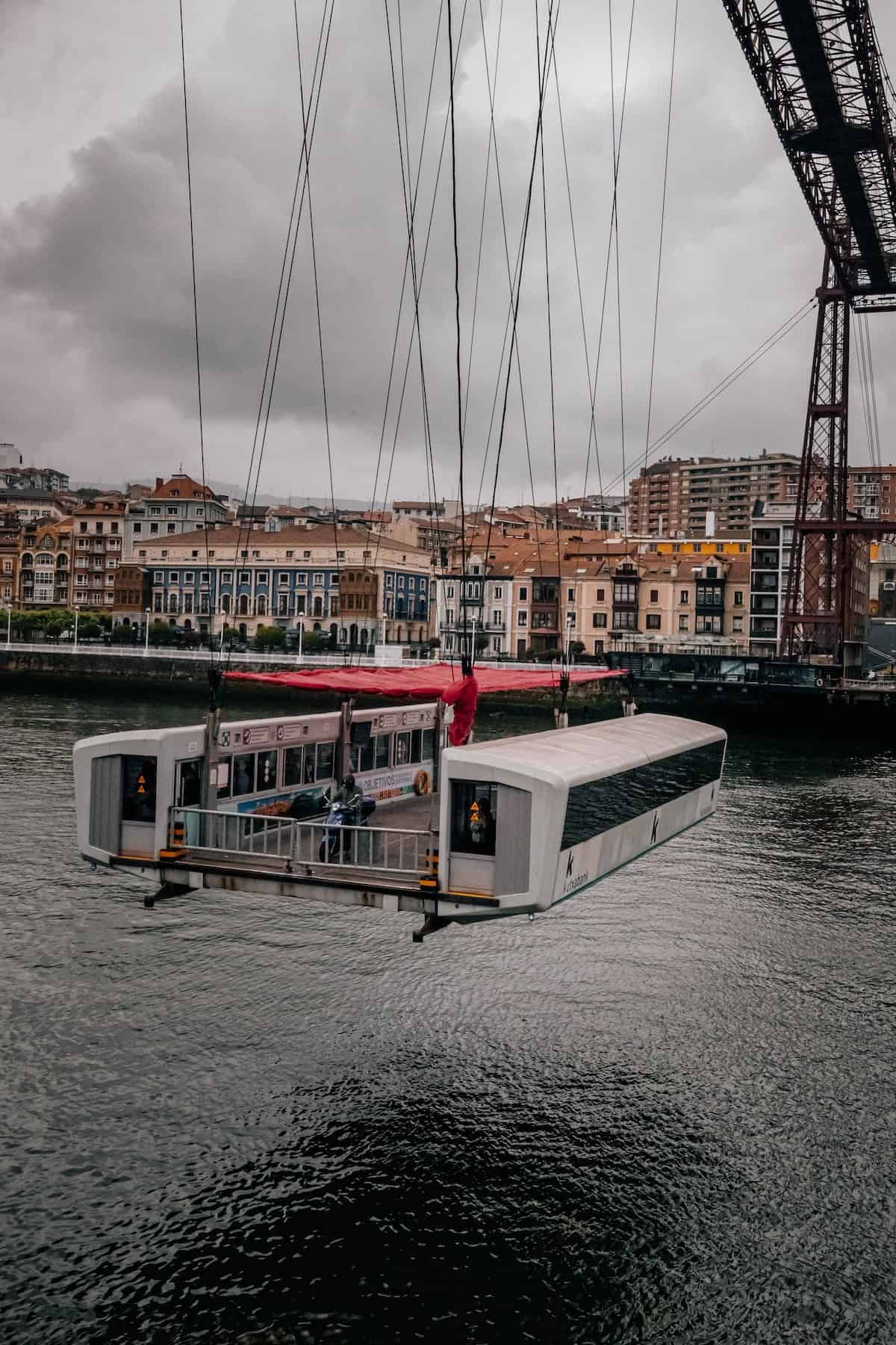 A suspended gondola is transporting a vehicle and passengers across a river, with an urban landscape and overcast sky in the background.