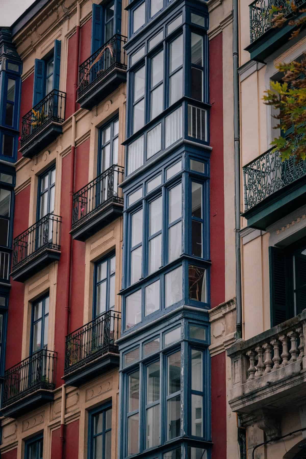 A multi-story building with red faรงade, black balconies, and large, vertically-stacked windows in blue frames. Nearby, there's a section with beige walls and green balconies.