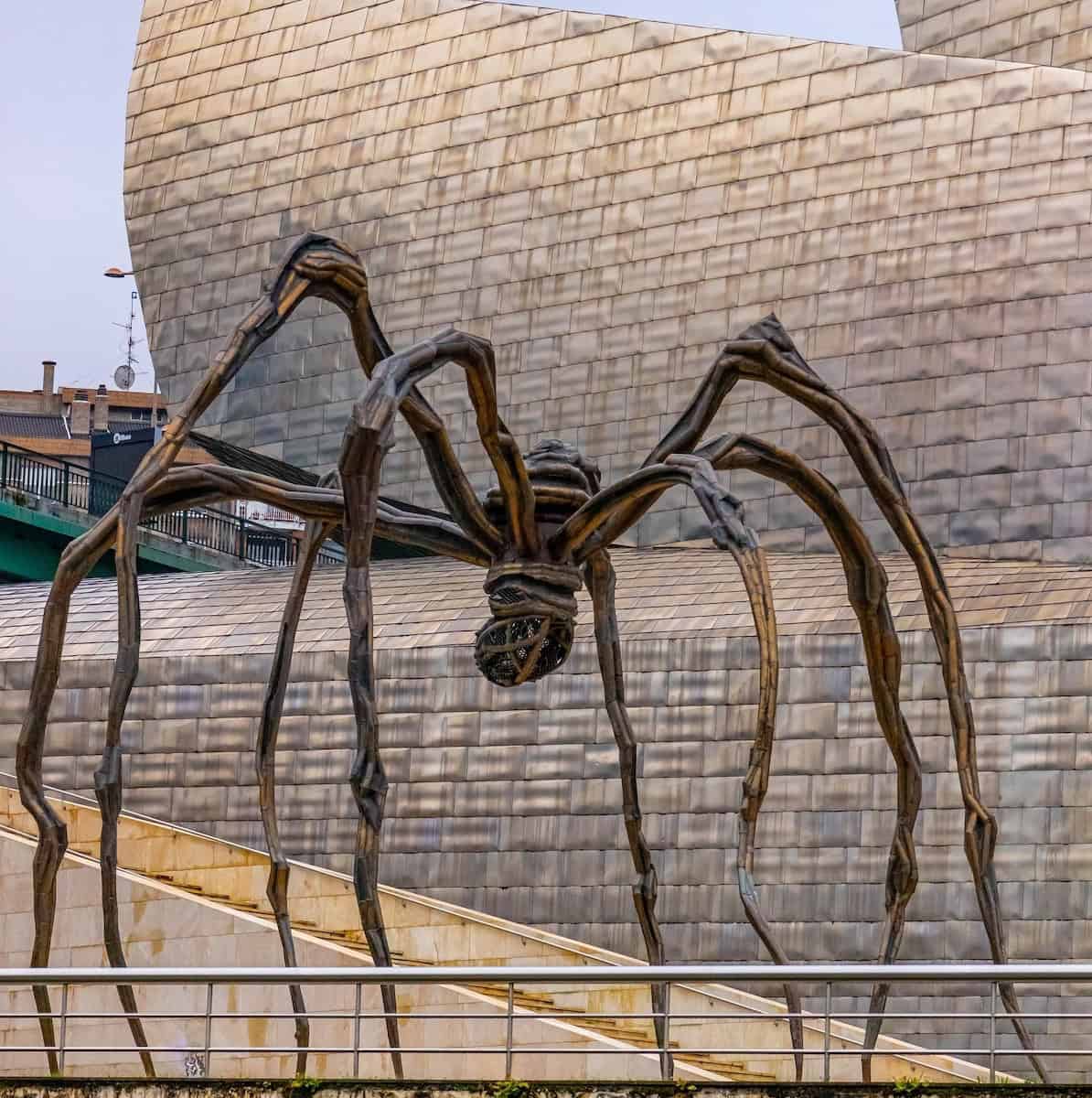 A large bronze spider sculpture stands outside a modern, metallic building in Bilbao, making it a must-see for any travel guide.