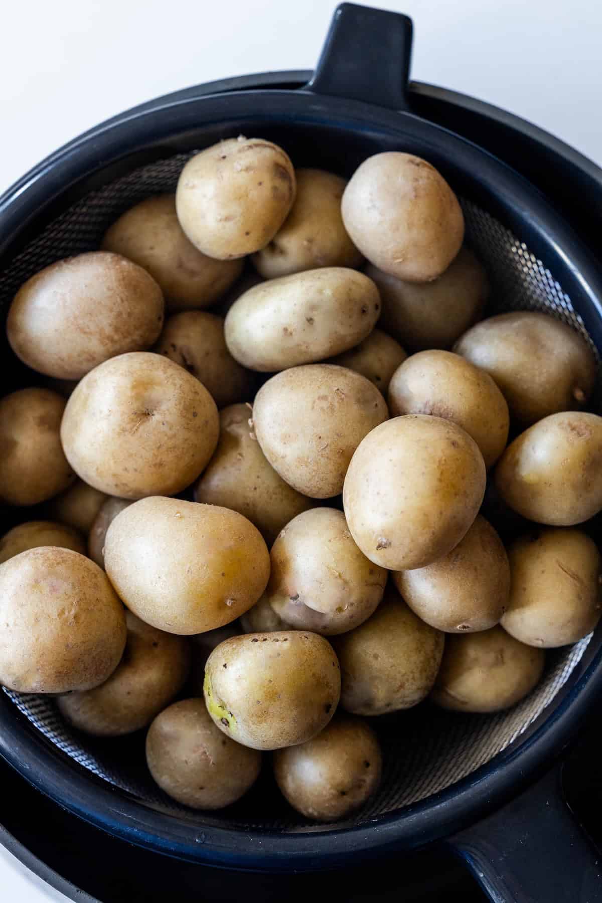 A metal colander filled with several small, unpeeled chive potatoes sits inside a black bowl on a white surface.