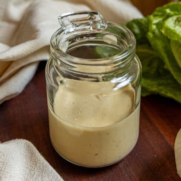A glass jar filled with creamy caesar dressing sits on a wooden surface next to a lettuce leaf and a light-colored cloth.