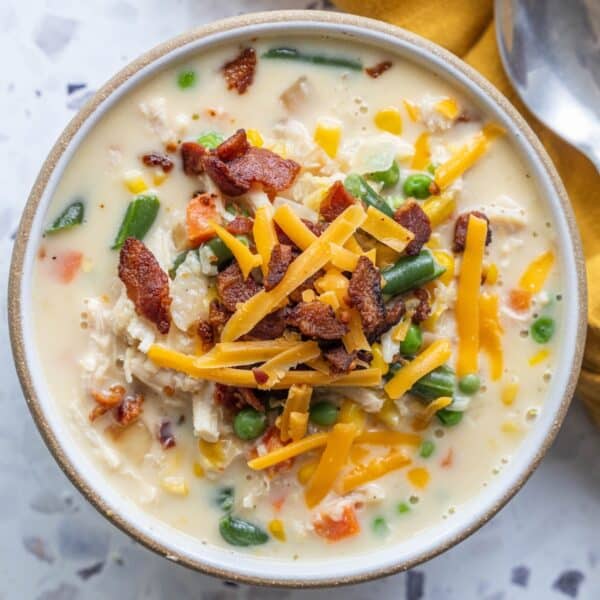 A bowl of creamy corn chowder topped with shredded cheddar cheese, crispy bacon, and filled with mixed vegetables like peas, green beans, carrots, and corn. A spoon and napkin are nearby.