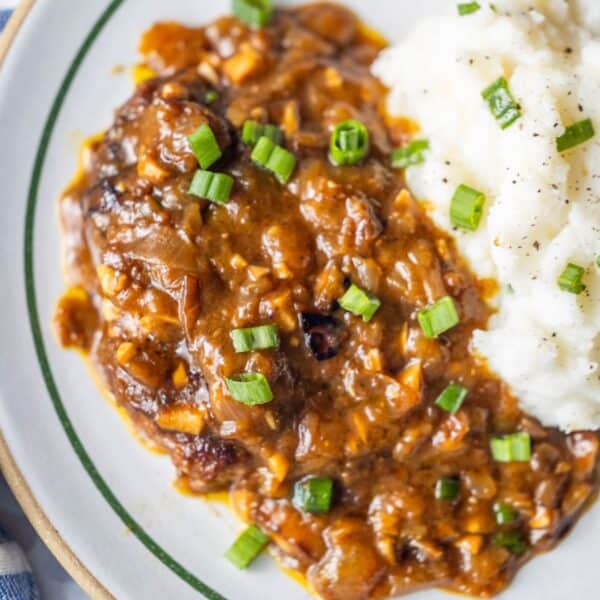 A plate with Salisbury steak, made using a classic country steak recipe, topped with brown onion gravy and served alongside mashed potatoes garnished with chopped green onions and black pepper.
