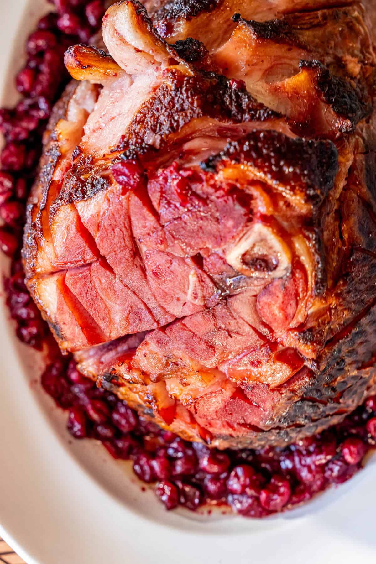 A Glazed Spiral Ham with a scored surface sits on a white plate, surrounded by a ring of cooked cranberries.