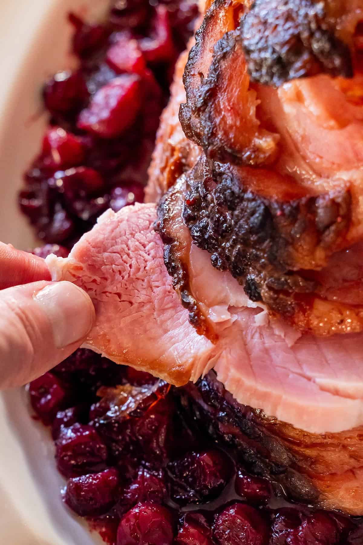 A hand pulls a slice of glazed spiral ham from a larger piece, served on a platter with a bed of cooked cranberries.