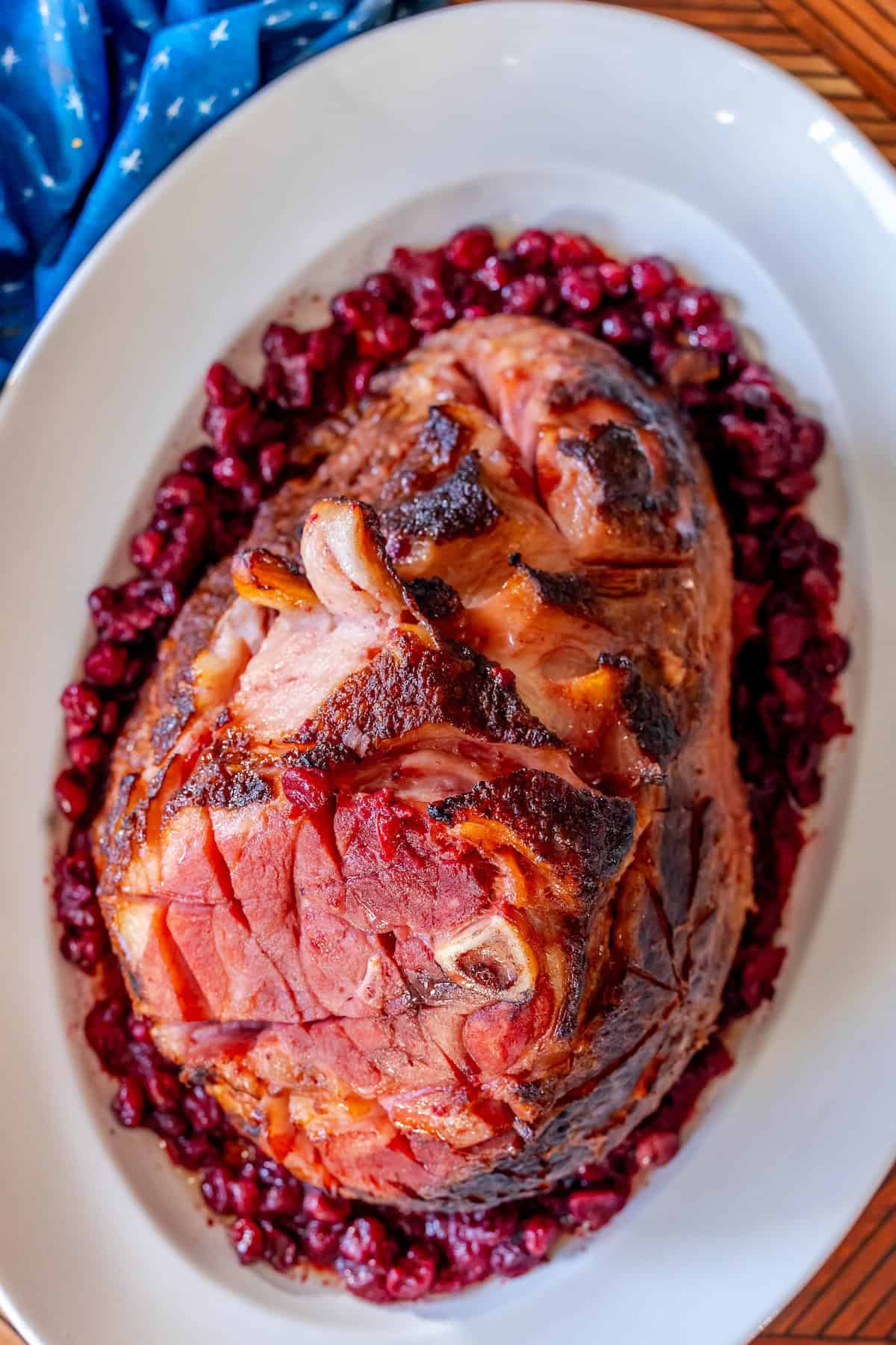 A glazed spiral ham with a browned, shiny surface is placed on an oval white platter, surrounded by a layer of cooked cranberries for a festive touch.