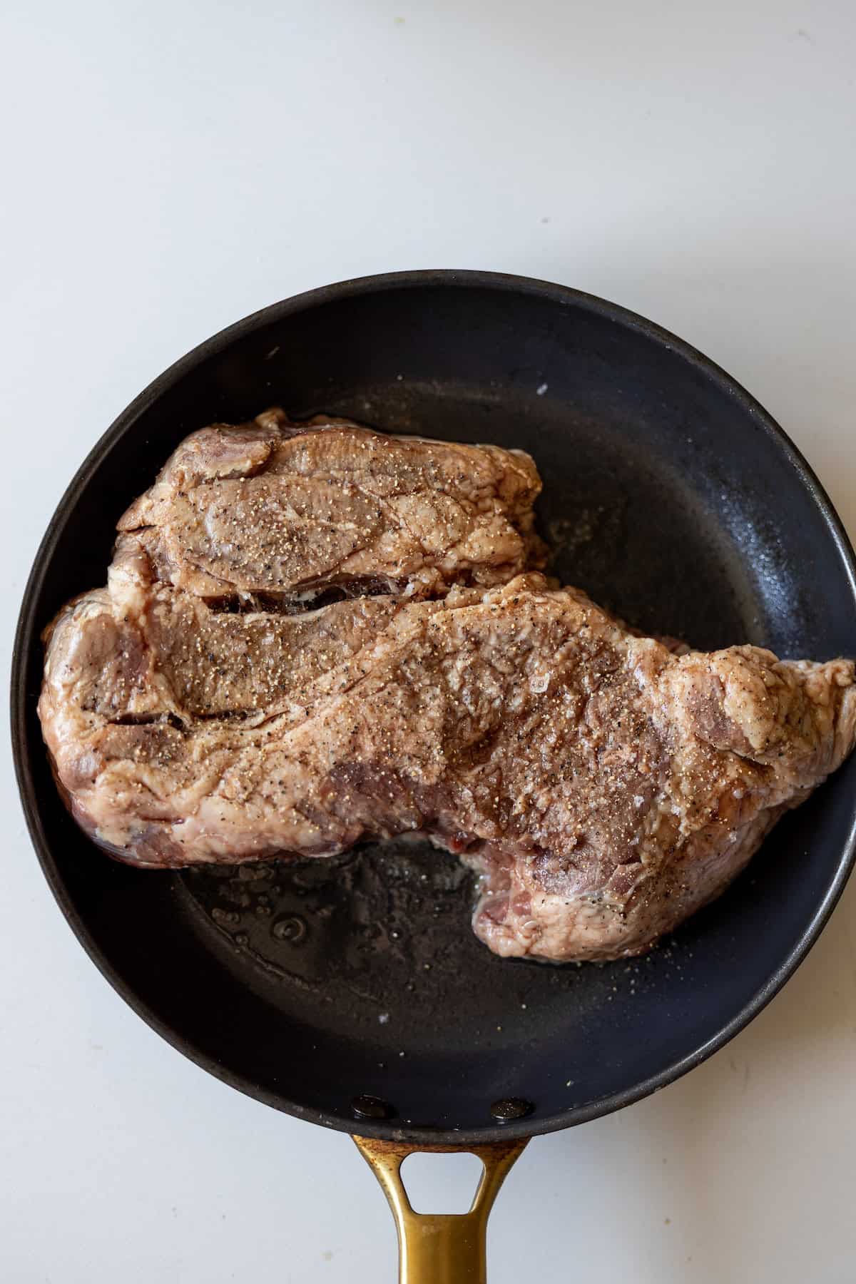A large, seasoned steak cooking in a black frying pan with a gold handle on a light-colored surface.