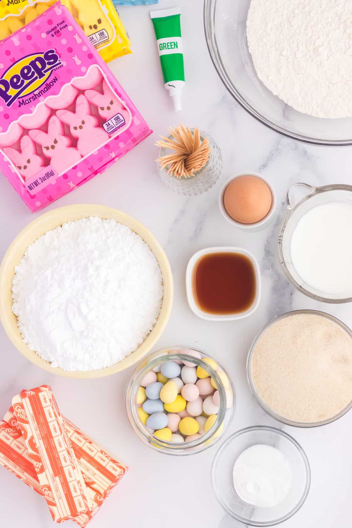 Baking ingredients on a countertop include flour, sugar, an egg, milk, vanilla, icing color, mini chocolate eggs, butter—and for a fun twist—Peeps marshmallows for delightful cupcakes.