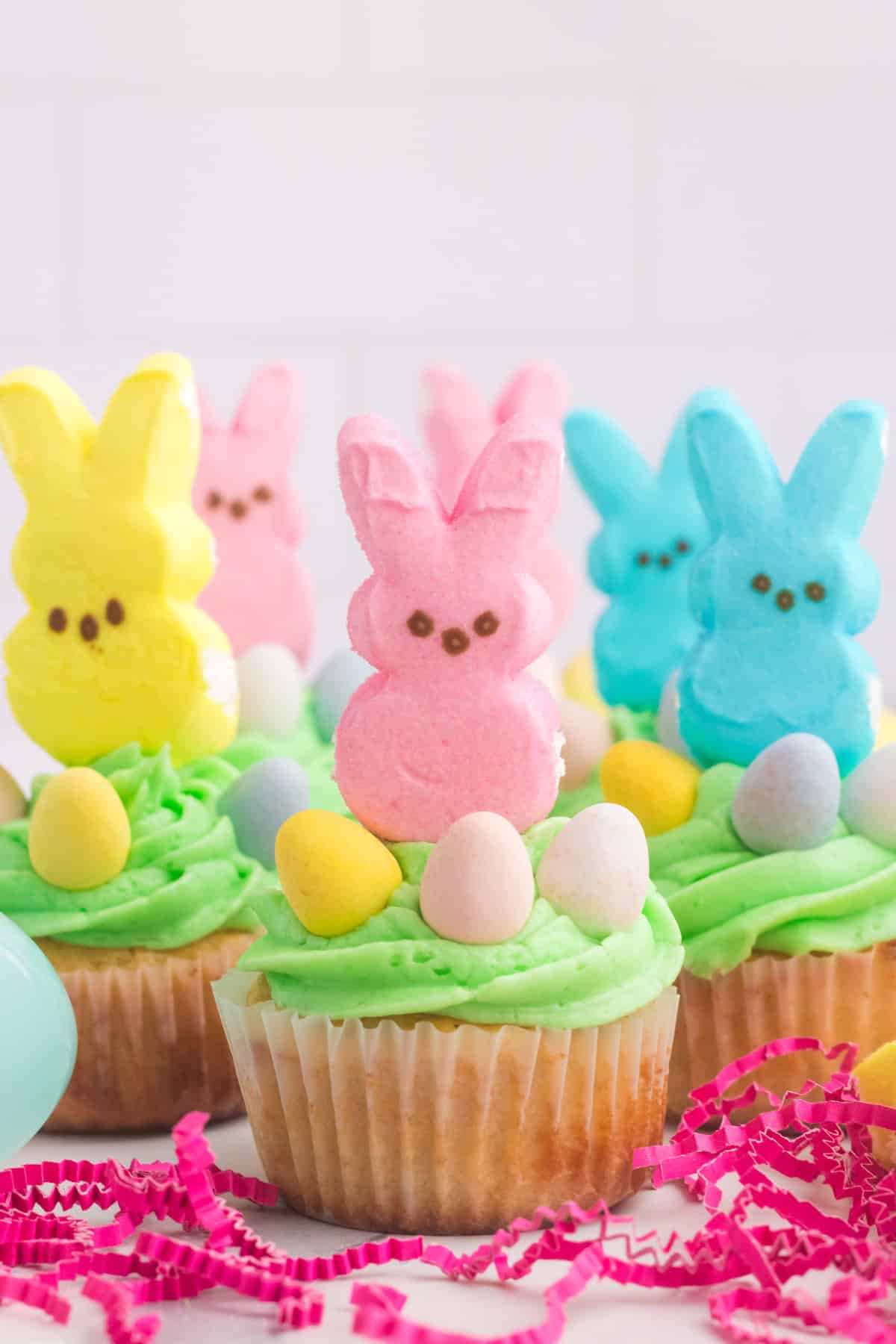 Cupcakes with green frosting, topped with colorful marshmallow Peeps and candy eggs, surrounded by pink shredded paper.