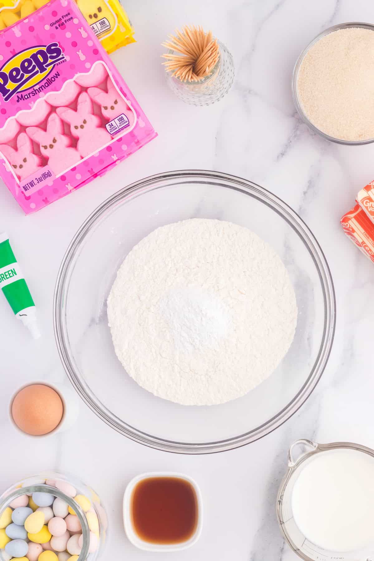 Baking essentials on a marble counter feature flour in a bowl, sugar, an egg, milk, vanilla extract, Peeps marshmallow chicks, green icing for cupcakes, butter, and colorful candies with toothpicks.