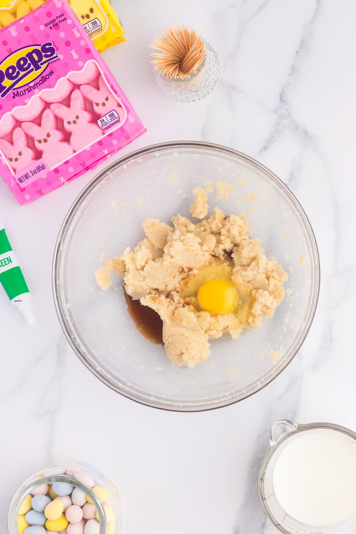 A mixing bowl contains cookie dough mixture with an egg and vanilla. Nearby, marshmallow Peeps, candy eggs, a cup of milk, green icing, and toothpicks sit ready to transform cupcakes on the marble countertop.