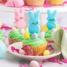 Cupcakes adorned with green frosting and marshmallow Peeps in pink, blue, and yellow. Pastel candy eggs nestle atop while decorative paper shreds and eggs surround the plate on a pristine white surface.