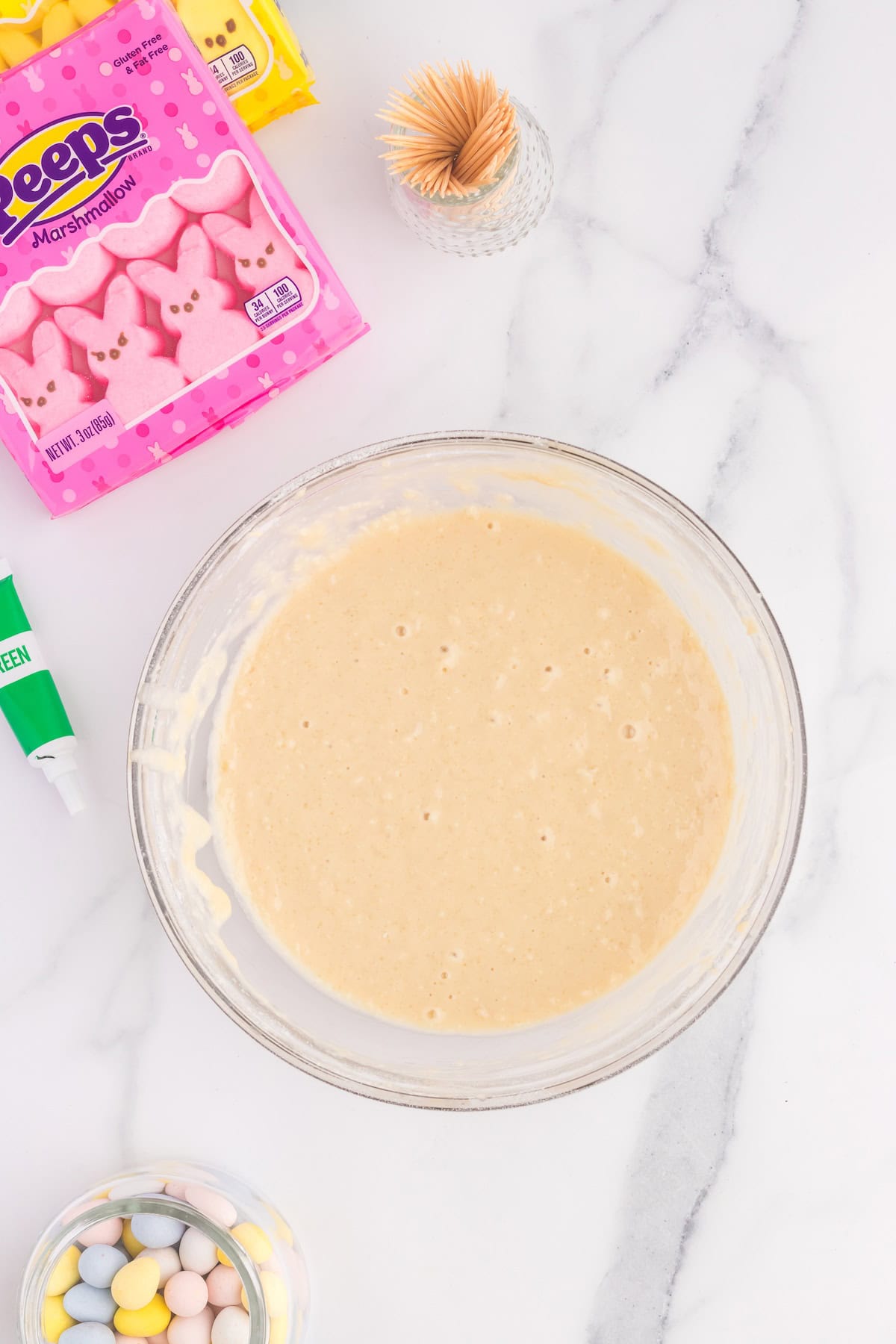 A mixing bowl with creamy batter is placed on a marble surface, ready to create delightful cupcakes. Nearby are pink marshmallow Peeps, pastel candy eggs, and a container with toothpicks waiting to decorate these sweet treats.