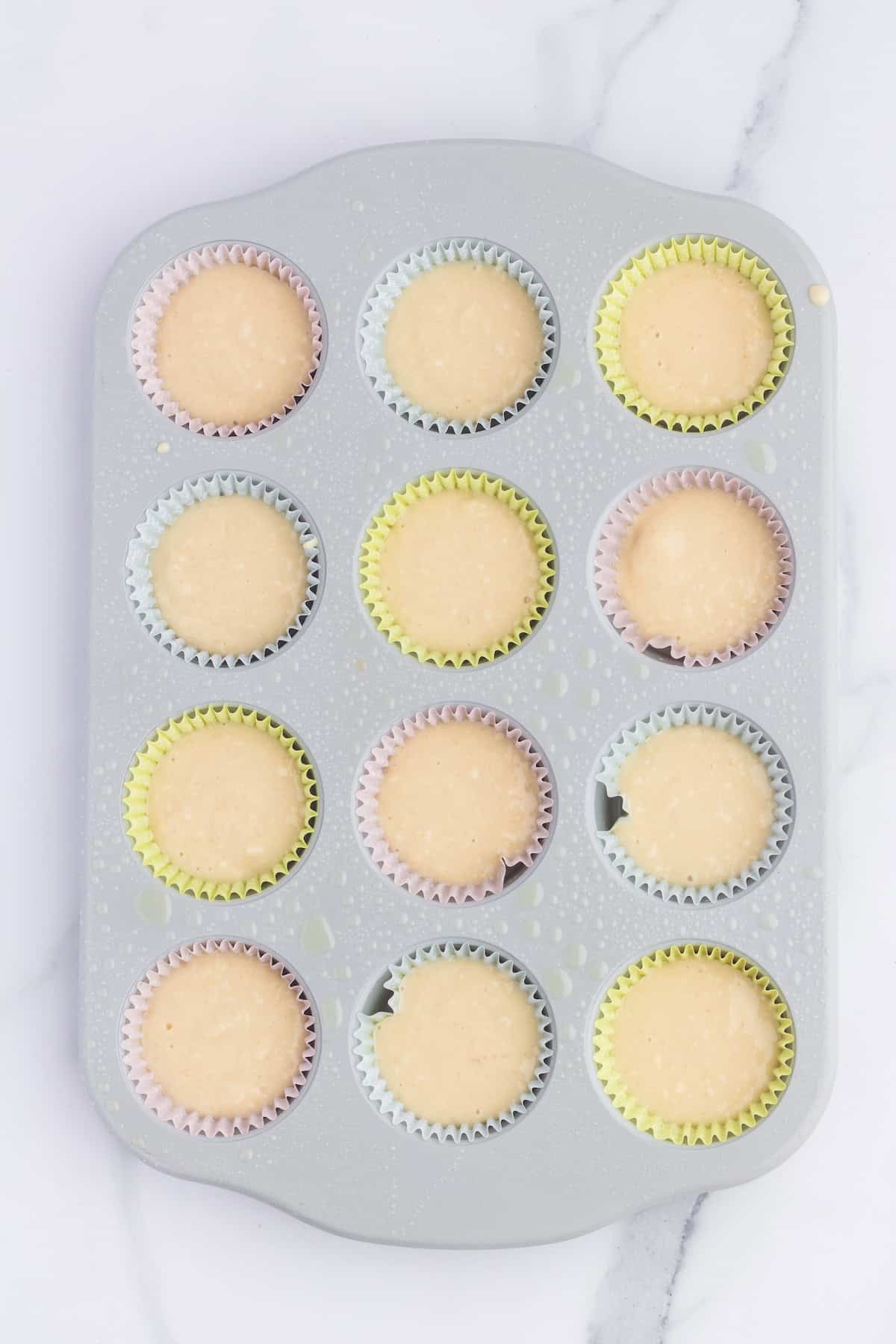 A muffin tray with twelve unbaked cupcakes, some topped with festive Peeps, sits on a marble counter in colorful paper liners.