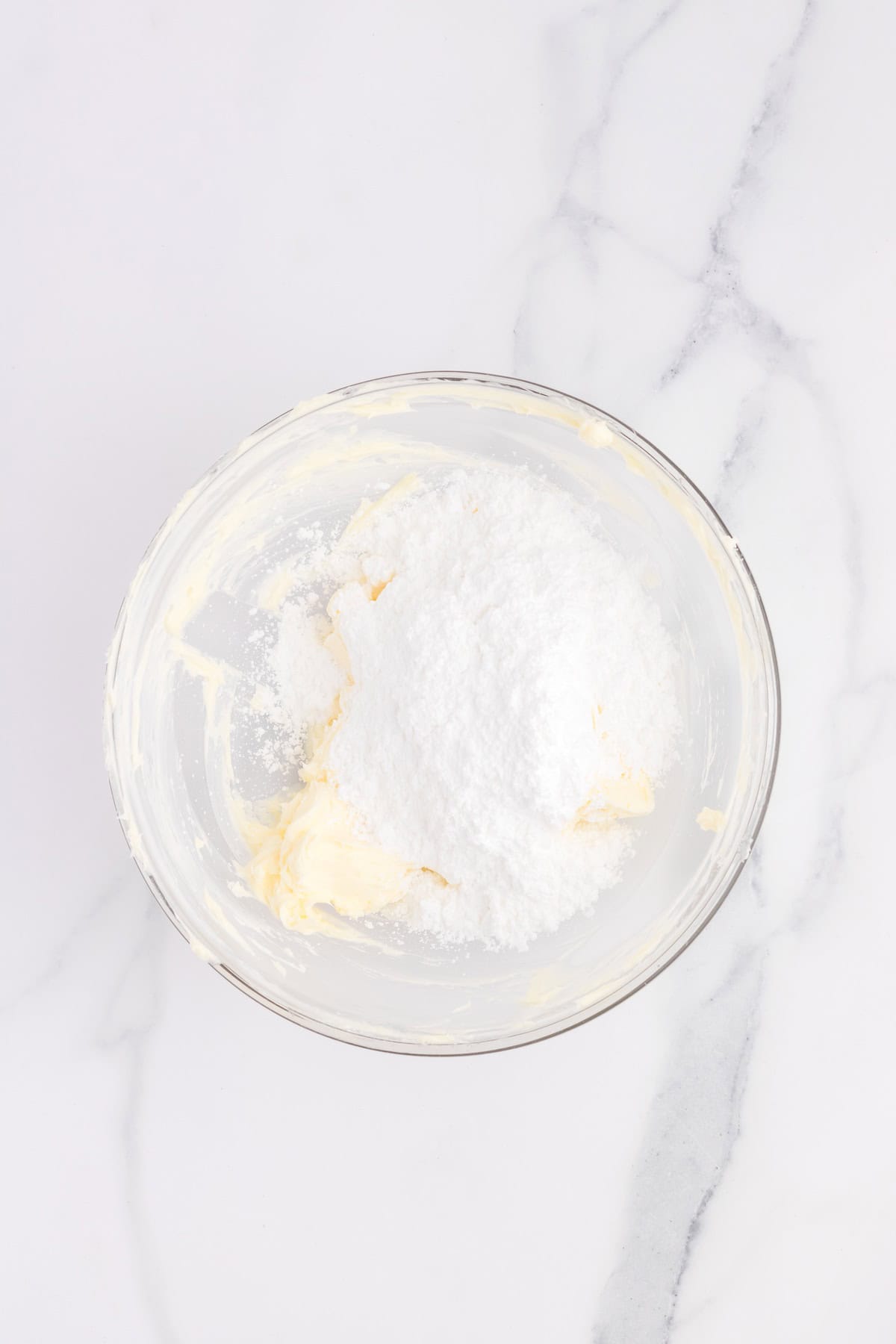 A glass bowl brimming with powdered sugar and cream sits gracefully on a white marble surface, ready to transform into the perfect topping for pastel-colored cupcakes.