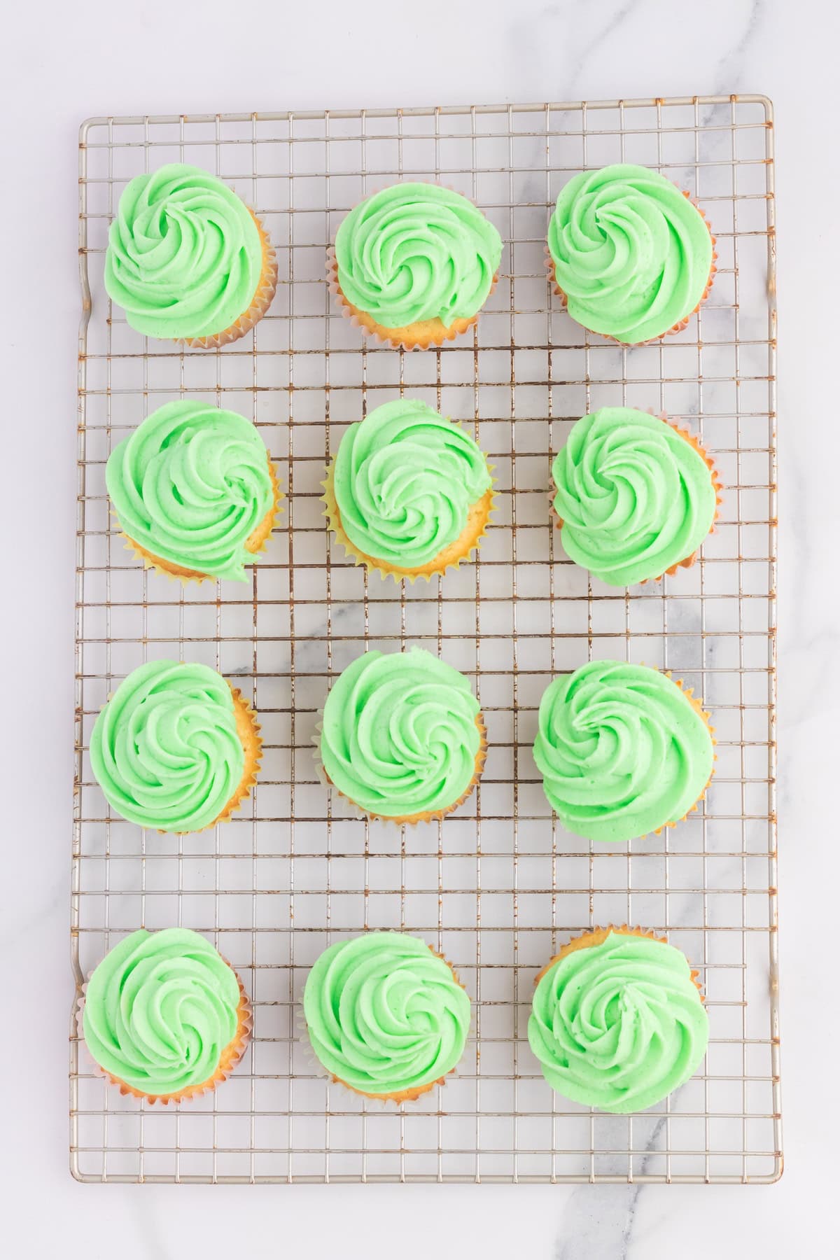 Twelve cupcakes with green swirled frosting topped with Peeps rest on a wire cooling rack, creating a delightful display on the marble surface.