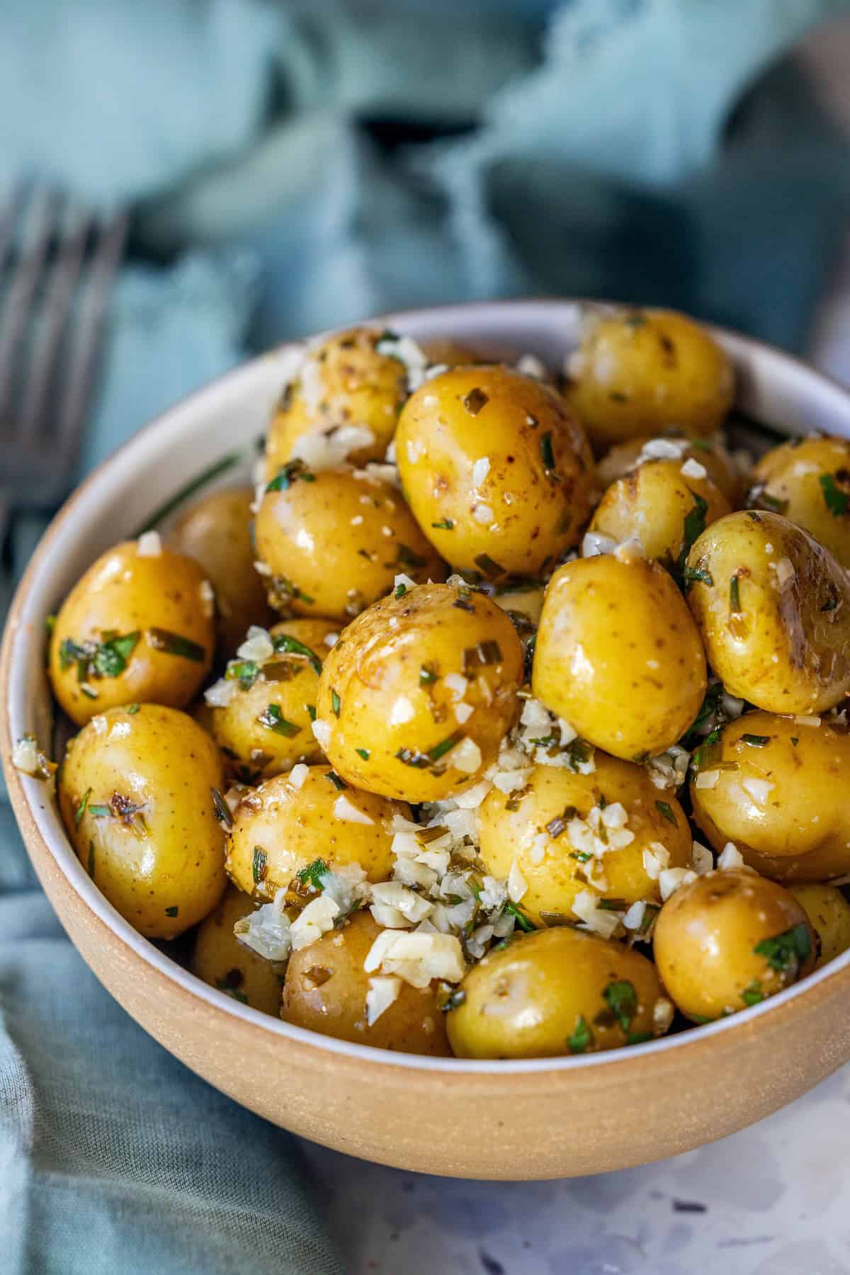 A bowl of garlic and chive potatoes, seasoned and garnished with chopped herbs and minced garlic, sits on a table with a blue cloth in the background.
