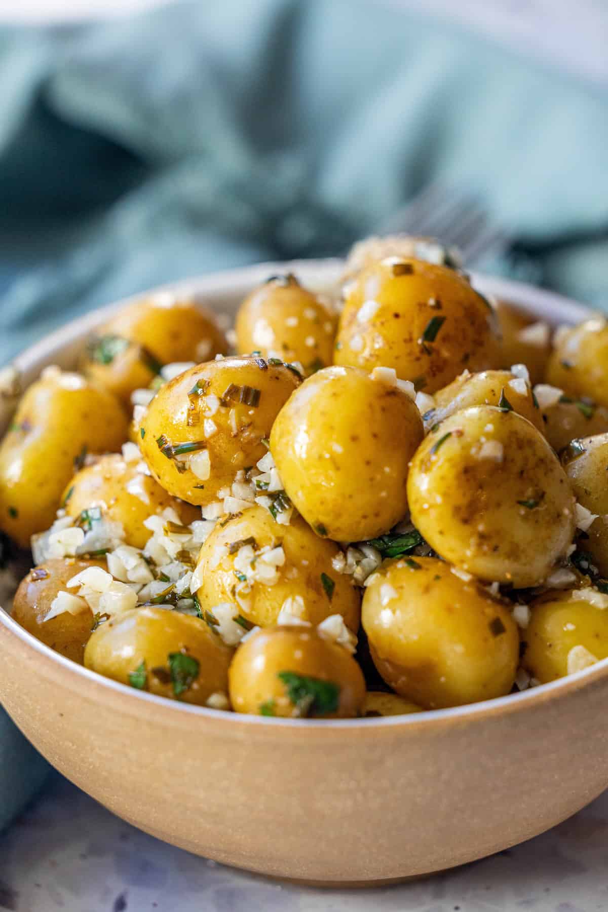 A bowl of garlic and chive potatoes garnished with chopped herbs and minced garlic sits on a table with a green napkin in the background.