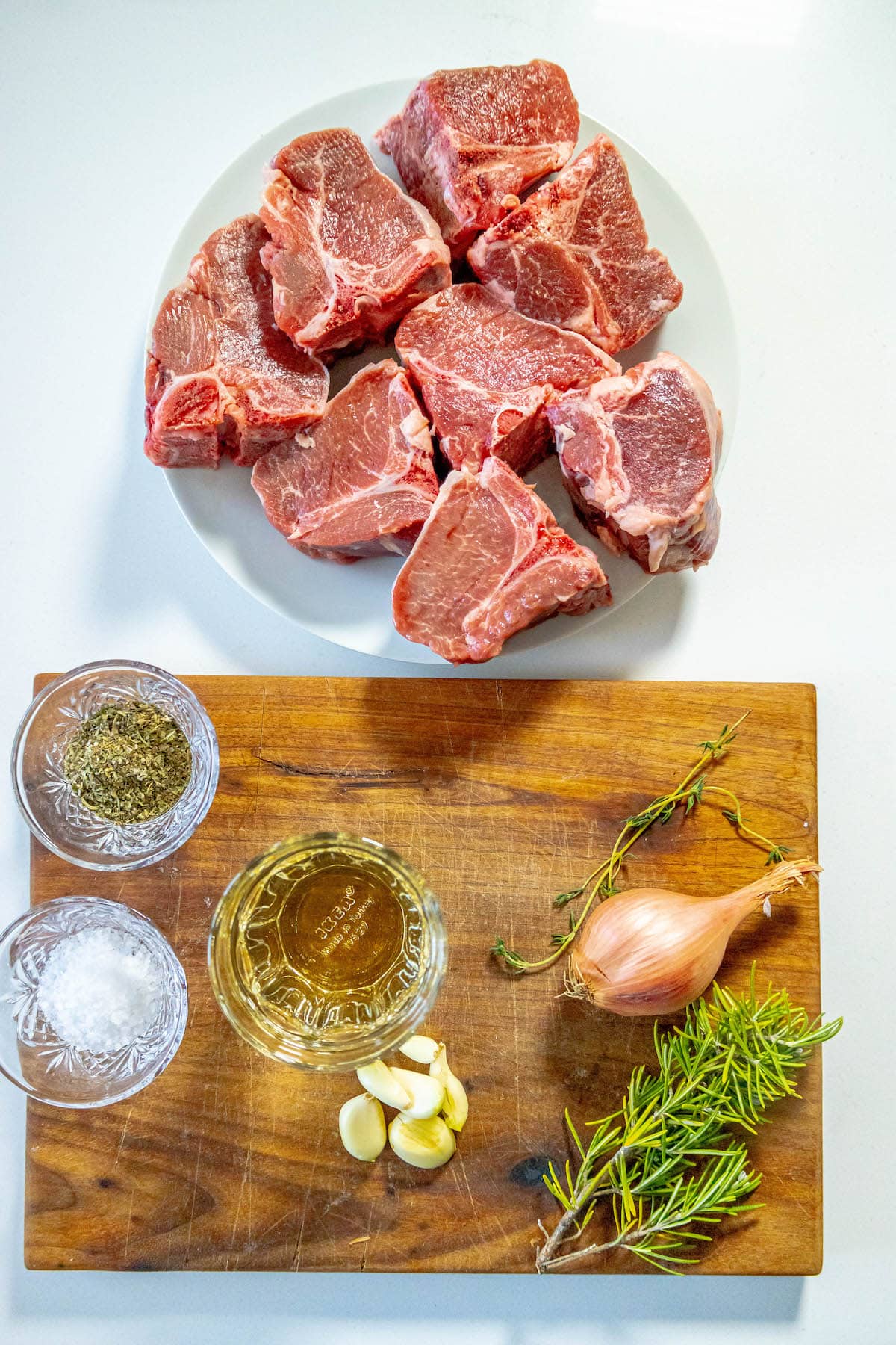 raw lamb chops on a white plate next to a cup of wine, herbs on a table, salt, a shallot, and fresh cracked pepper