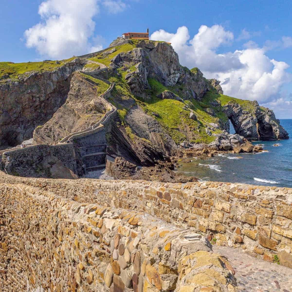 A stone path winds its way up to a small building atop a steep rock formation overlooking the sea under a partly cloudy skyโa must-see when you visit Basque Country.