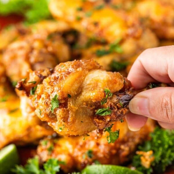A hand holding a piece of crispy fried chicken garnished with herbs, hinting at the flavor of garlic wings, with more pieces and lime wedges in the background.