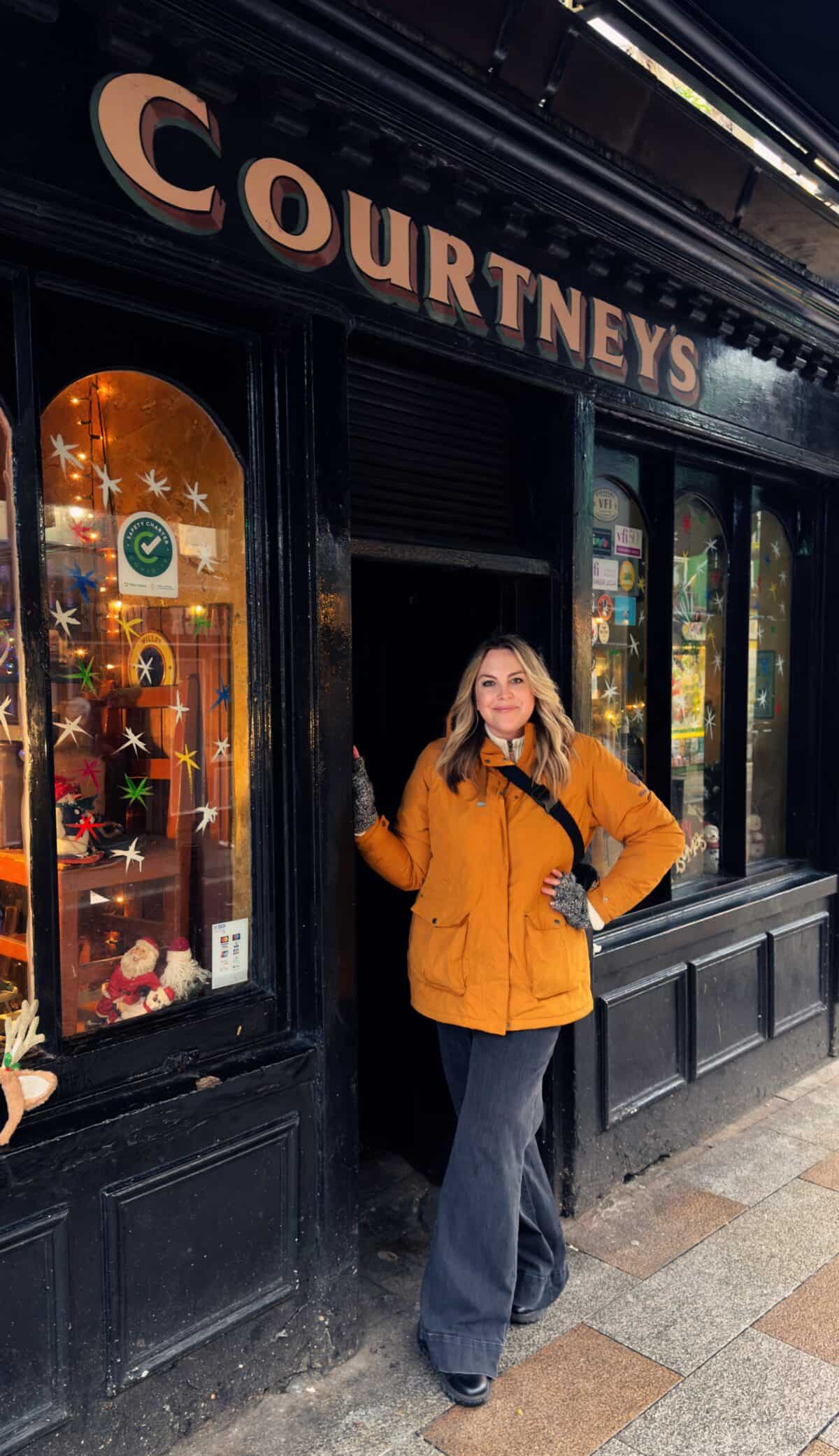 A person in a yellow jacket stands in the doorway of a black storefront labeled "Courtneys," reminiscent of Courtney O'Dell Sweet C's Designs, with decorated windows displaying various items.