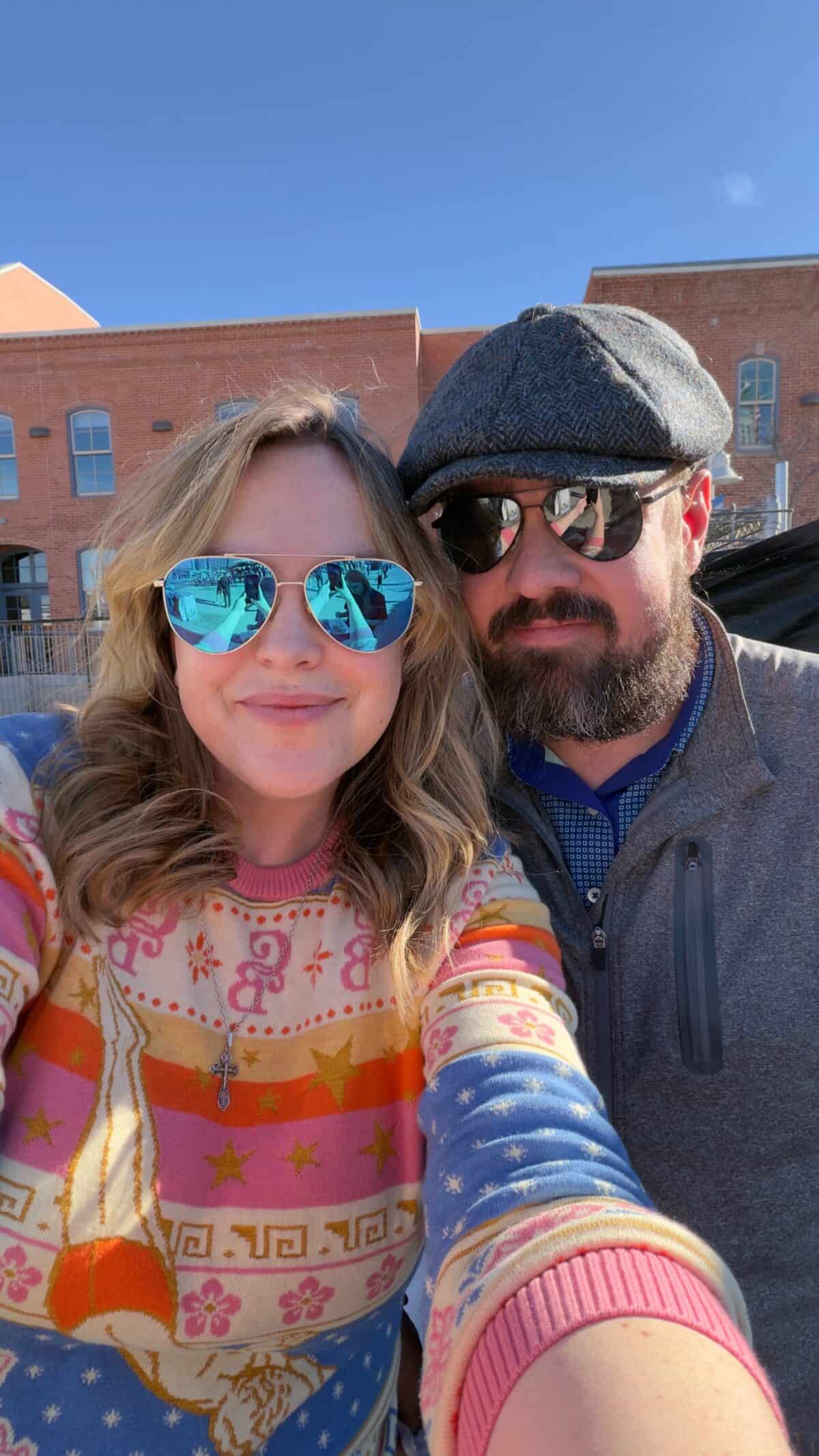 Two people wearing sunglasses pose for a selfie outdoors on a sunny day, with brick buildings in the backgroundโcapturing a fun moment for the Sweet C's Designs Blog.