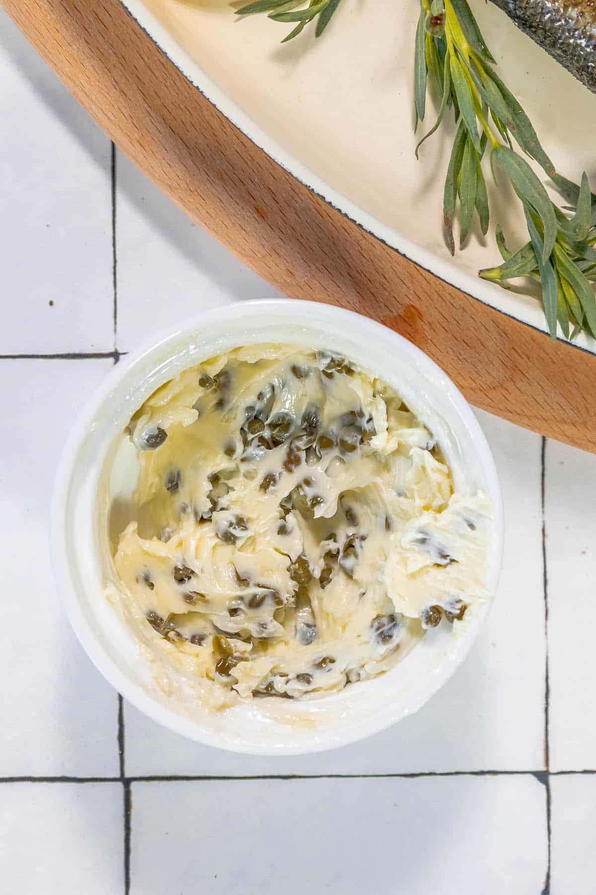 A bowl of lemon caper butter and a bowl of roasted branzino on a tiled floor.