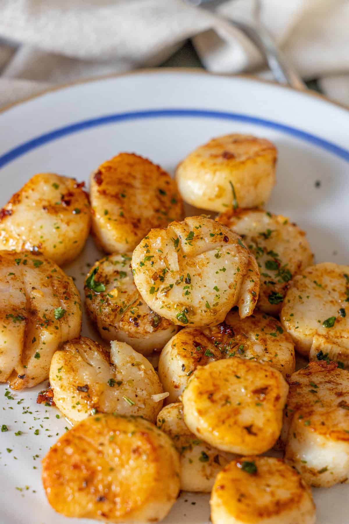 A white plate with Perfect Pan Seared Scallops, garnished with chopped herbs, set on a table with a fork and napkin in the background.