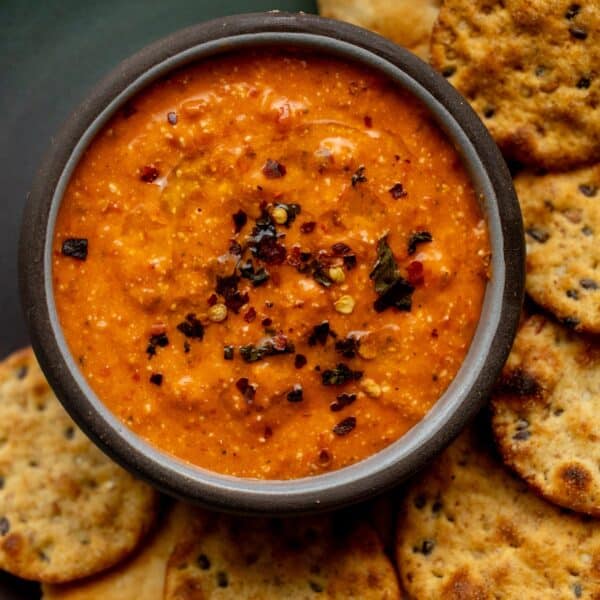 A bowl of red pepper dip topped with dried herbs and chili flakes, surrounded by round, seeded crackers.