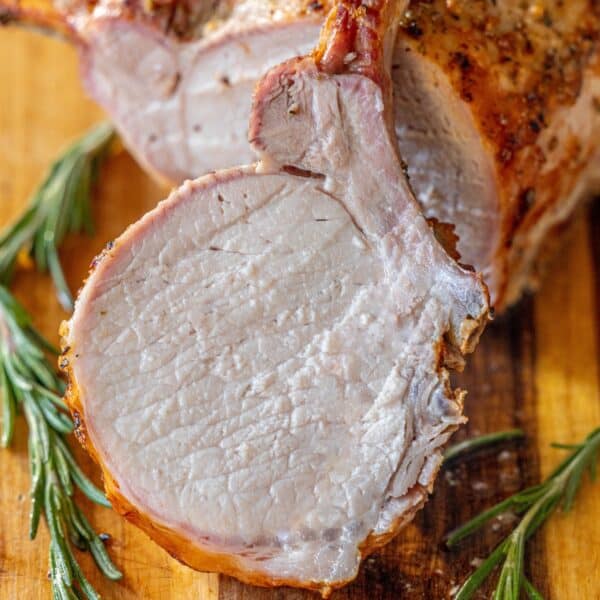 A roasted rack of pork, sliced to reveal its juicy interior, is displayed on a wooden board with fresh rosemary sprigsโa true showcase of the best roasted pork.