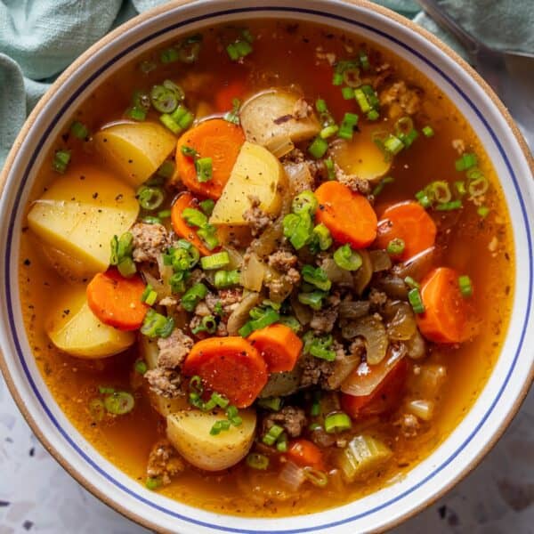 A bowl of sausage vegetable soup with ground meat, potato chunks, carrot slices, celery, onions, and chopped green onions in a clear broth.