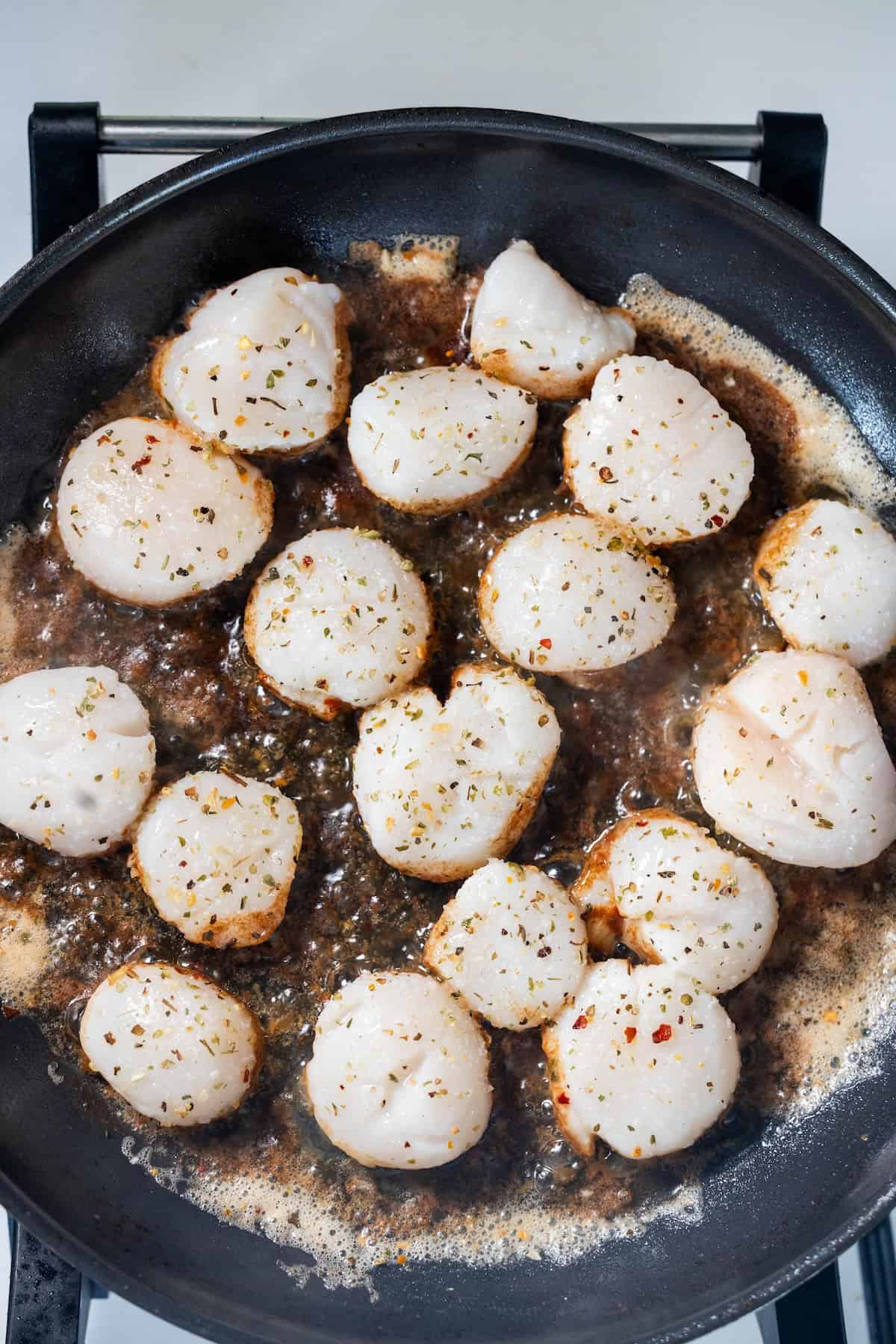 Perfect Pan Seared Scallops cooking in a frying pan with bubbling sauce, sprinkled with fresh herbs and spices.