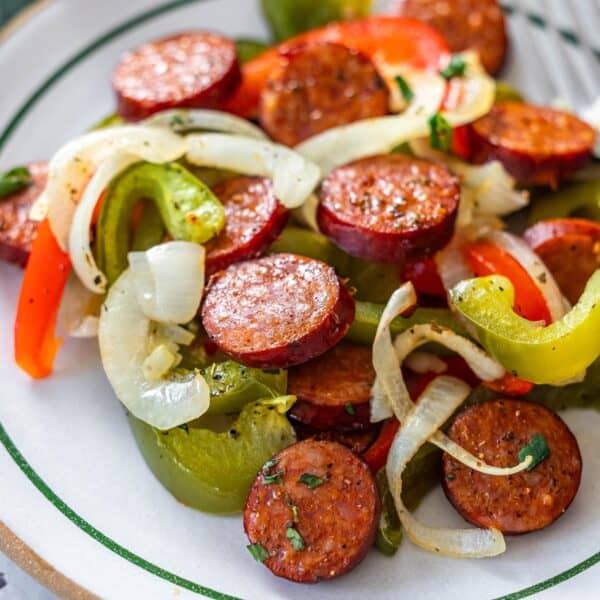 A plate of sliced sausage, sautรฉed onions, and bell peppers, seasoned with herbs and pepper, features this classic Sheetpan Kielbasa and Peppers dinner, served on a white dish with green trim.