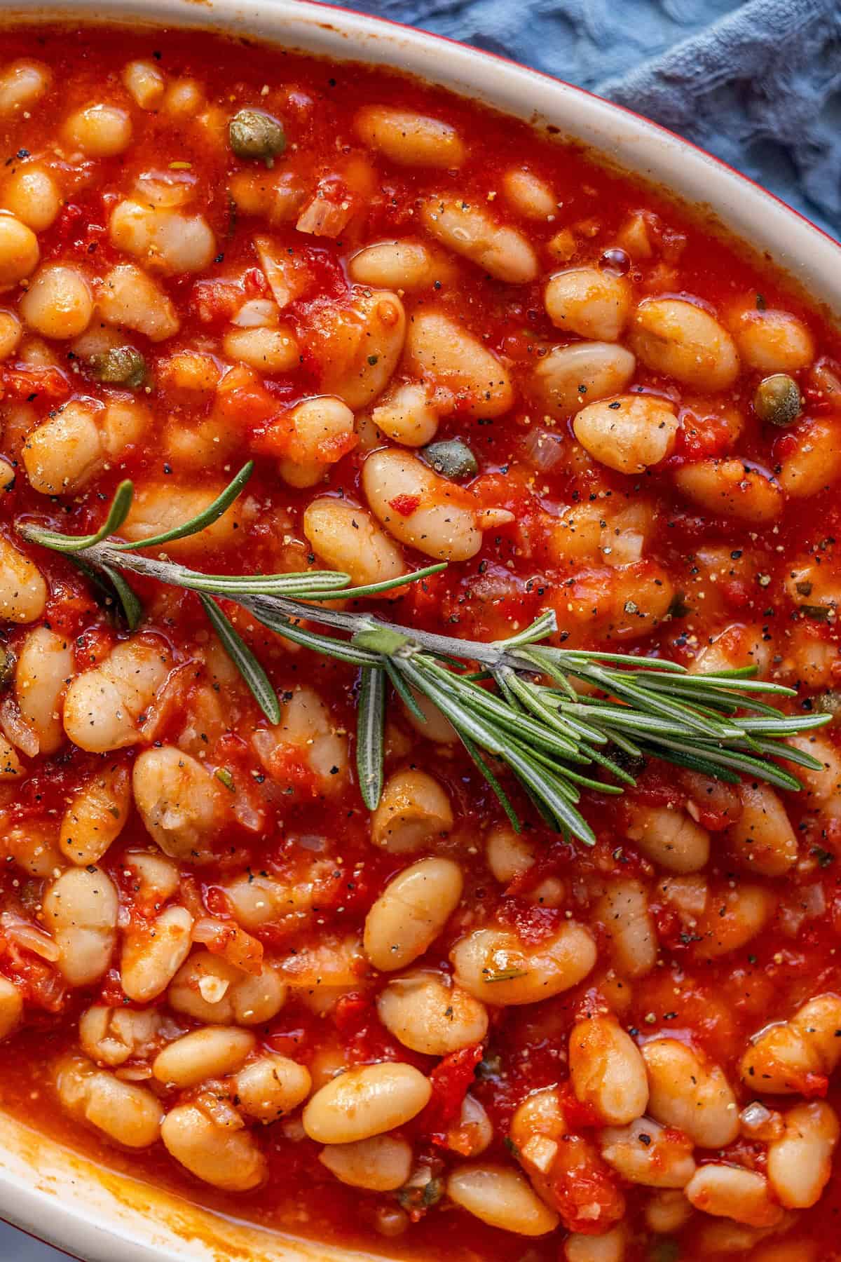 Close-up of a dish with white beans in tomato sauce, garnished with a sprig of rosemary.