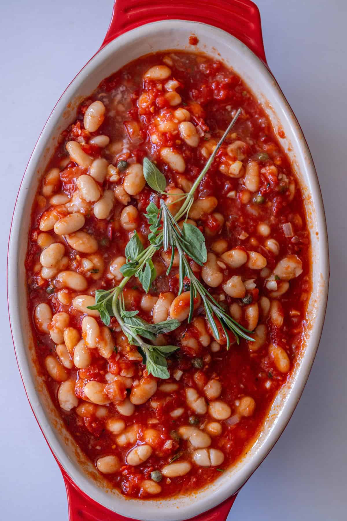 An oval red baking dish filled with Tuscan Roasted White Beans in tomato sauce, garnished with fresh herbs.
