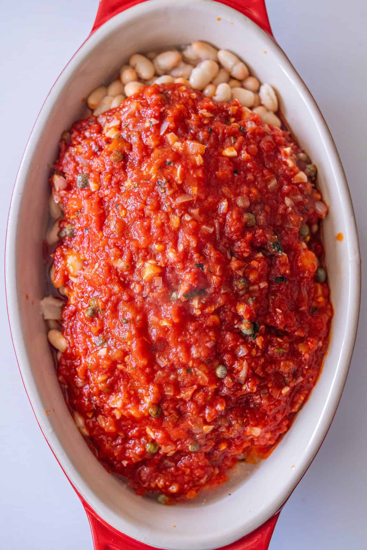 An oval baking dish filled with Tuscan Roasted White Beans and topped with a layer of chunky tomato sauce.