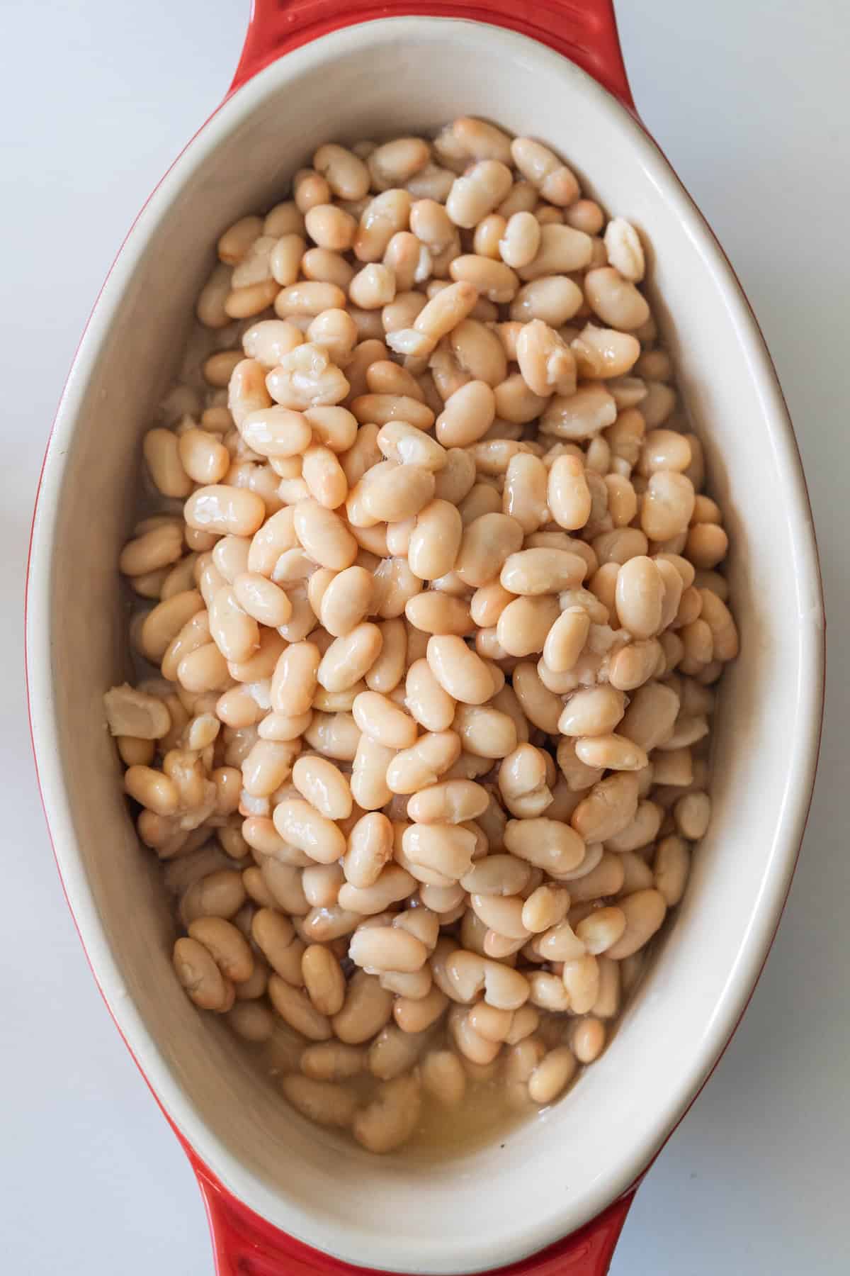 An oval dish filled with Tuscan roasted white beans in savory liquid, placed on a white surface.