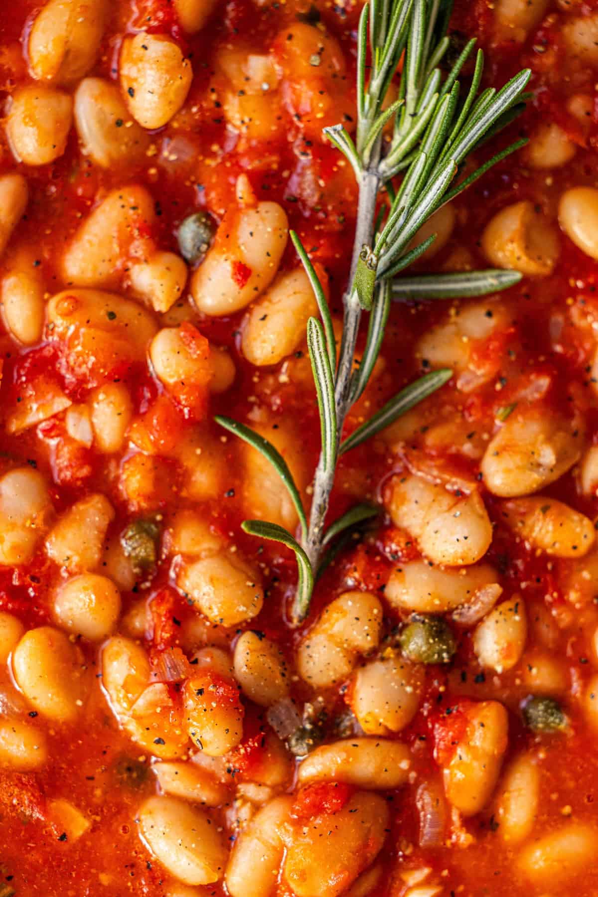 Close-up of white beans in a tomato-based sauce, garnished with a sprig of fresh rosemary and sprinkled with black pepper.
