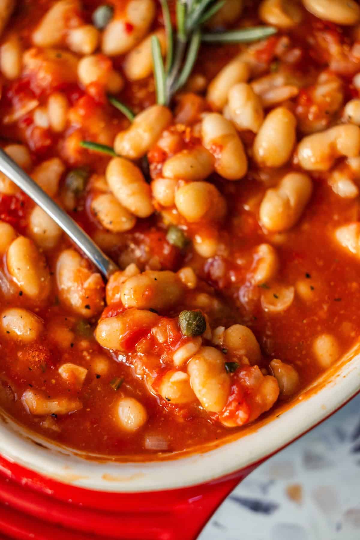 Close-up of a spoon in a dish of stewed white beans in tomato sauce, garnished with rosemary.