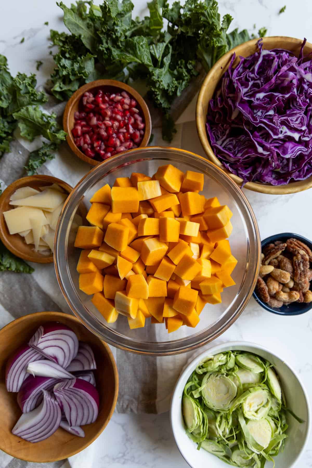 A bowl of cubed butternut squash is surrounded by bowls of sliced red cabbage, brussels sprouts, pomegranate seeds, cheese slices, nuts, and kale—perfect for creating a vibrant winter salad on a white surface.