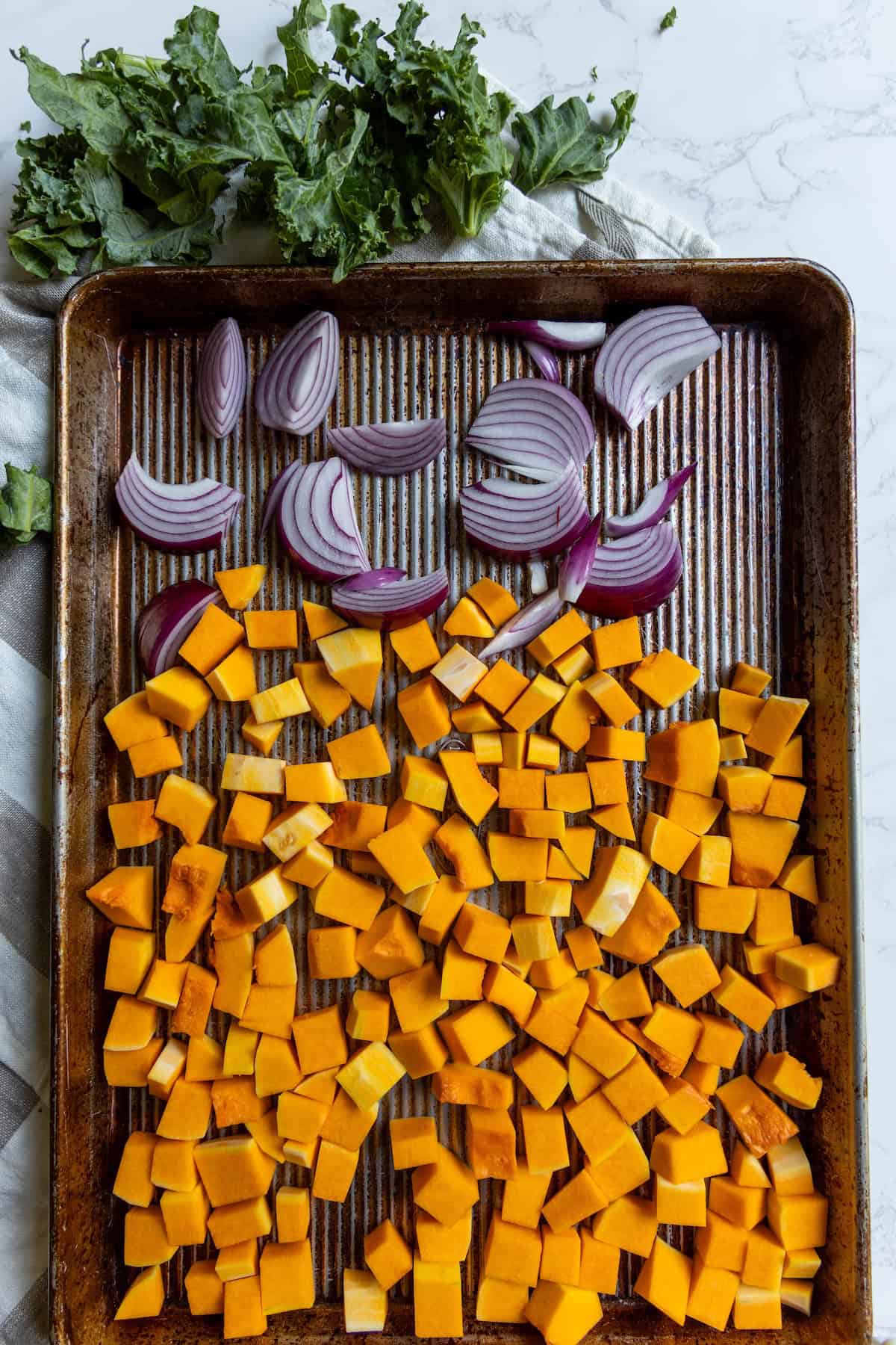 A baking sheet with diced butternut squash and sliced red onions, with chopped kale on the side—perfect for preparing a hearty winter salad.